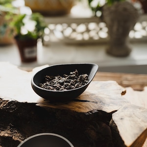 May include: A small, matte black, teardrop-shaped bowl filled with dark, tightly rolled dried tea leaves. The bowl has a smooth, curved rim and sits atop a piece of natural, light-colored wood with a rough, textured bark edge.