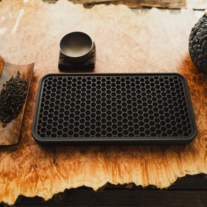 Puede incluir: Un juego de té dispuesto sobre una superficie de madera. El juego incluye una tetera negra con tapa, una taza de té pequeña, una bandeja de madera con hojas de té sueltas, una bandeja negra con un patrón de panal y una maceta texturizada con una planta.