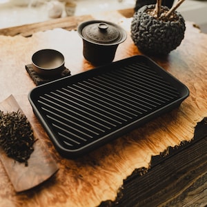 Peut inclure: Un service à thé noir disposé sur une table en bois. L'ensemble comprend un plateau noir, un petit bol, une théière couverte et une cuillère en bois avec des feuilles de thé en vrac. Une sphère texturée est également présente.