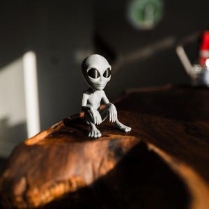 May include: A small, gray alien figurine with large black eyes sits on a dark wooden surface. The figurine is in a seated position, with its arms resting on its knees. The background is blurred, with sunlight casting shadows.
