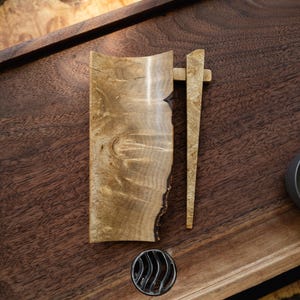 May include: A handcrafted wooden vessel with a natural edge, accompanied by a matching wooden tool. The vessel and tool are light brown with unique wood grain patterns. A small, dark metal object is also present, all set on a dark wooden surface.