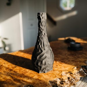 May include: A dark gray, textured vase with a unique, organic design. The vase has a narrow neck and a wider base, sitting on a wooden table. The vase's surface has a carved, irregular pattern.