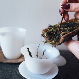 Może przedstawiać: Osoba wlewa luźną zieloną herbatę z drewnianej łopatki do białego ceramicznego kubka do herbaty. Kubek do herbaty stoi na białym spodeczku. Liście herbaty są zielone i brązowe.