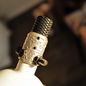 May include: Close-up of a decorative lamp base. The lamp features a white, rounded base with a silver-colored, patterned metal section. The top of the lamp has a black, woven-textured shade. The lamp is designed for indoor use.
