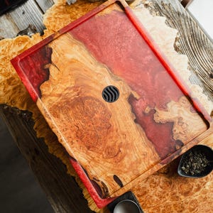 May include: A rectangular serving tray crafted from natural light brown burl wood and translucent red resin. The tray features a smooth, polished surface with a central circular black drain insert and a raised border along the edges.
