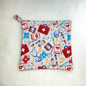 May include: A red and white quilted hot pad with a medical theme. The hot pad is covered in a pattern of red first aid kits, bandages, pills, and other medical supplies. The hot pad has a loop handle.