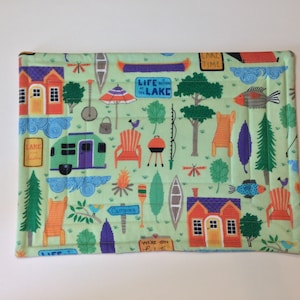 May include: A green fabric placemat with a camping theme. It features illustrations of cabins, a camper, trees, fish, and a canoe. The text on the placemat reads "Life is better at the lake" and "Lake Time".