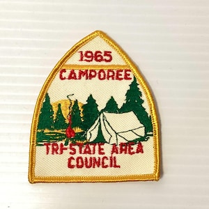 May include: A shield-shaped embroidered patch with a yellow border. It features a white background with a red campfire, green pine trees, and a white tent. The text reads 1965 CAMPOREE and TRI-STATE AREA COUNCIL in red embroidered lettering.
