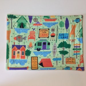 Puede incluir: Un artículo de tela rectangular verde claro con un tema de camping. El diseño incluye ilustraciones de una tienda de campaña, una casa, peces, árboles y una caravana. El texto incluye "Welcome to the Lake" y "Life at the Lake".