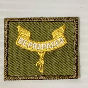 May include: A square embroidered patch with an olive green background and a dark brown border. Centered on the patch is a yellow ribbon graphic with white text that reads BE PREPARED, featuring a small yellow rope knot hanging below the ribbon.