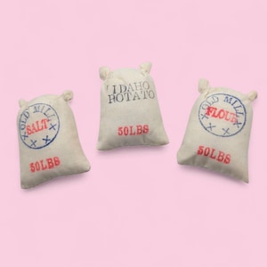 May include: Three off-white fabric sacks with stitched details and ear-like handles. One sack reads "SALT" in a blue circle, another "IDAHO POTATO", and the third "FLOUR". All sacks are labeled "50 LBS" in red.