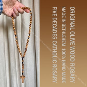 May include: A brown wooden Catholic rosary with five decades, made in Bethlehem. The rosary is made of olive wood and has a black cord. The text on the image reads "Original Olive Wood Rosary, Made in Bethlehem 100% Hand Made, Five Decades Catholic Rosary".