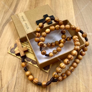 May include: A wooden rosary with brown beads and a brown cord. The rosary is in a brown box with the words "Hand made" printed on the side. The box is on a wooden surface.
