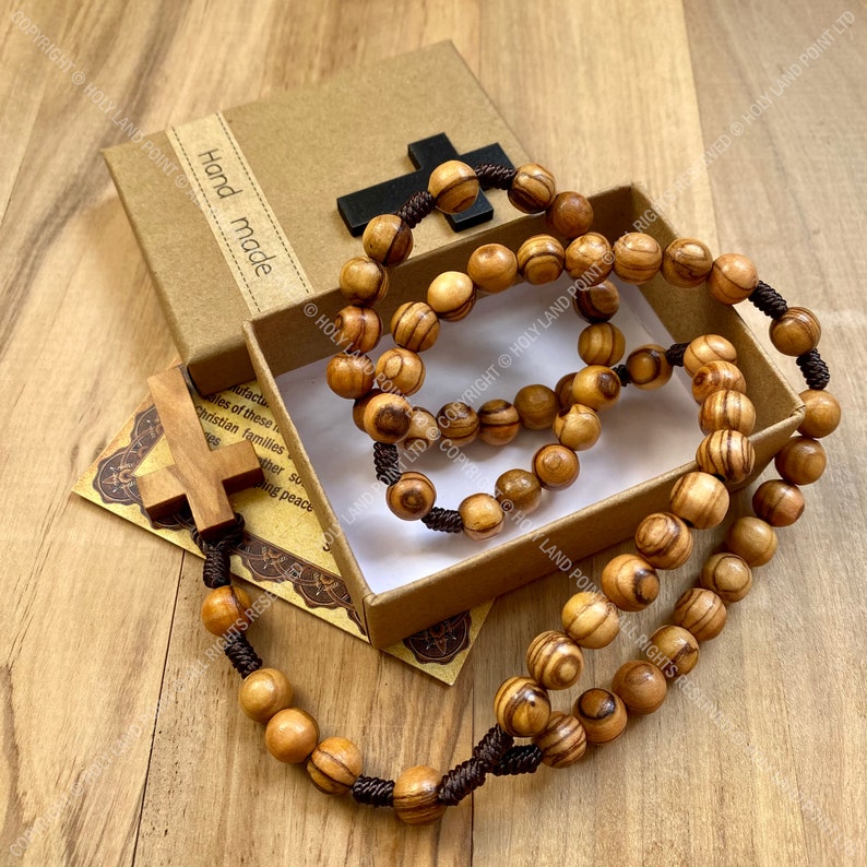 May include: A wooden rosary with brown beads and a brown cord. The rosary is in a brown box with the words "Hand made" printed on the side. The box is on a wooden surface.