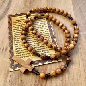 May include: A brown wooden rosary with a brown cord and a wooden cross. The rosary beads are made of olive wood from the Holy Land. The card in the background has a certificate of conformity and information about the olive wood.