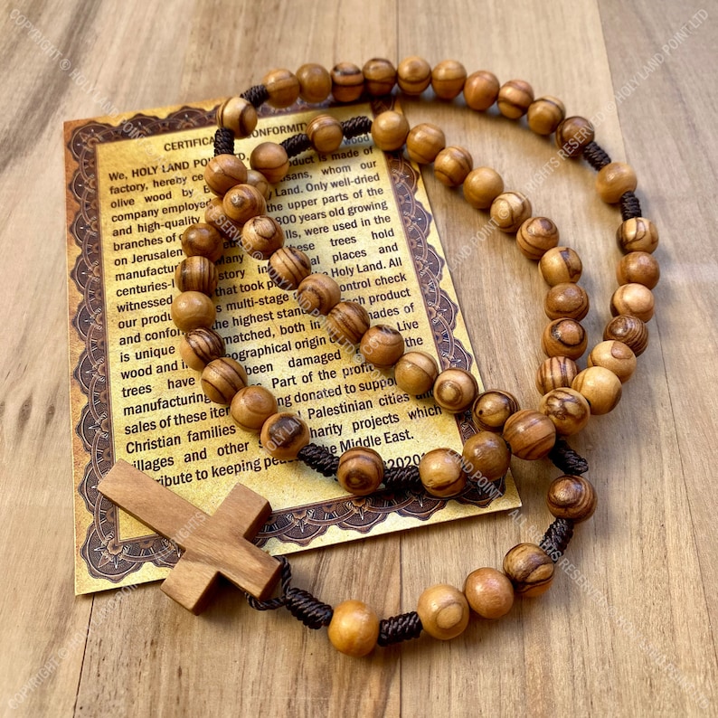 May include: A brown wooden rosary with a brown cord and a wooden cross. The rosary beads are made of olive wood from the Holy Land. The card in the background has a certificate of conformity and information about the olive wood.