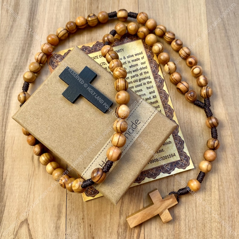 May include: A brown wooden rosary with a dark brown wooden cross. The rosary is on a wooden surface and comes in a brown box with a certificate of authenticity. The certificate reads "Hand Made" and "Rights Reserved Holy Land Point Ltd."
