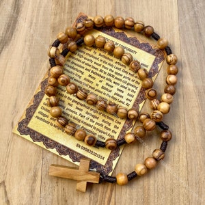 May include: Olive wood rosary beads with a brown cord and a wooden cross. The beads are brown with darker brown stripes. The rosary is on a certificate of conformity that reads "I hereby certify that this olive wood..." and includes the serial number SN 090820201554.