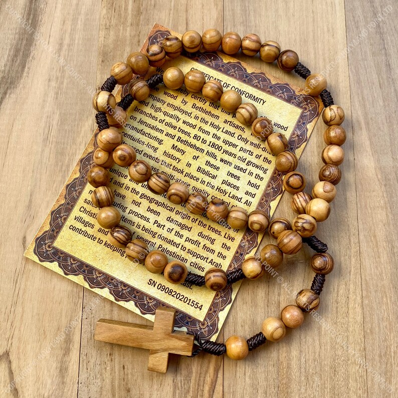 May include: Olive wood rosary beads with a brown cord and a wooden cross. The beads are brown with darker brown stripes. The rosary is on a certificate of conformity that reads "I hereby certify that this olive wood..." and includes the serial number SN 090820201554.