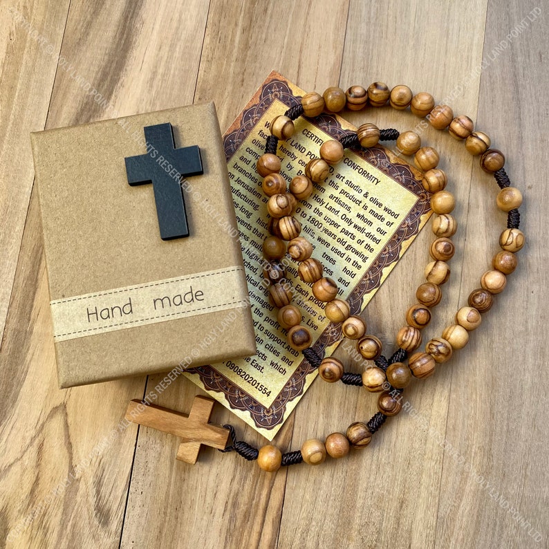 May include: A brown wooden box with the words "Hand Made" on the front. The box contains a wooden rosary with brown beads and a small wooden cross. A certificate of authenticity is included, which states that the olive wood is from the Holy Land and is 1800 years old.