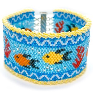 May include: A beaded bracelet with a blue, white, and yellow design featuring orange and yellow fish. The bracelet has a silver clasp.