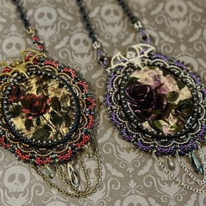 May include: Two oval pendants on chains, each featuring a central rose image and a metal bat accent at the top. The left pendant is framed with red and black beads, while the right pendant is framed with purple and black beads, both with dangling chains.