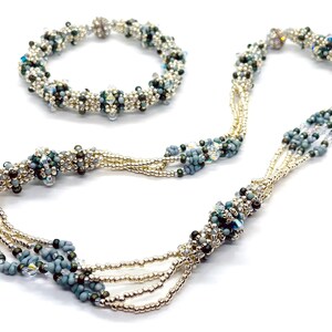 Puede incluir: Un conjunto de collar y pulsera de cuentas plateadas y azules. El collar es de múltiples hilos y presenta un diseño trenzado. La pulsera es de un solo hilo y también presenta un diseño trenzado.