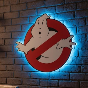 May include: A Ghostbusters-themed wall light featuring the iconic no-ghost logo with a white ghost figure inside a red circle with a red slash. The light has a blue neon glow and is mounted on a brick wall.