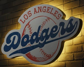 Könnte beinhalten: Ein Schild des Baseballteams Los Angeles Dodgers im Used-Look. Das Schild zeigt das Logo des Teams mit den Worten "Los Angeles" in Rot und "Dodgers" in Blau, vor einer Baseball-Grafik. Das Schild ist vor einer Backsteinmauer beleuchtet.