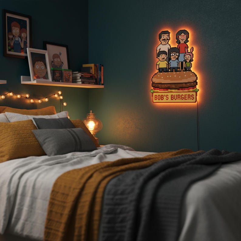 May include: A pixelated LED sign featuring the Bob's Burgers family atop a burger, with the text "BOB'S BURGERS" below. The sign emits a warm orange glow, mounted on a teal wall above a bed with yellow and gray pillows and a mustard-colored blanket.
