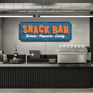 May include: A retro-style sign with the text "Snack Bar" in orange letters on a blue background. The sign also says "Drinks, Popcorn, Candy" in white letters. The sign is hanging above a black counter with a silver espresso machine and a black sign that says "Order Here".