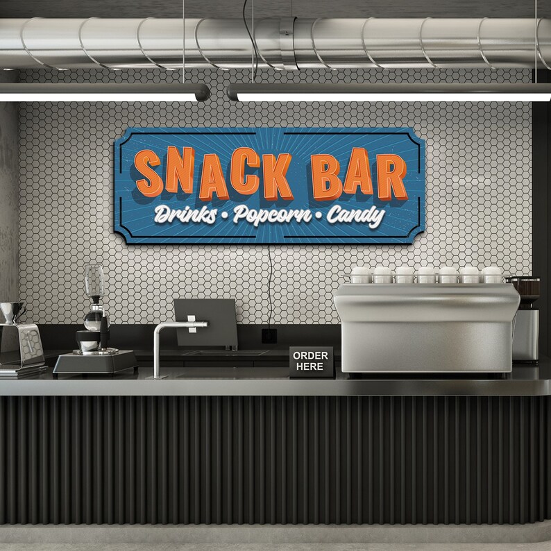 May include: A retro-style sign with the text "Snack Bar" in orange letters on a blue background. The sign also says "Drinks, Popcorn, Candy" in white letters. The sign is hanging above a black counter with a silver espresso machine and a black sign that says "Order Here".