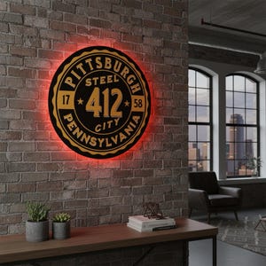 Op de afbeelding: Een ronde zwarte en gouden Pittsburgh-bord met de tekst "Pittsburgh Steel 412 City Pennsylvania" en een rode neon gloed. Het bord is bevestigd aan een bakstenen muur.