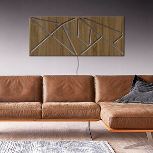 May include: A modern wooden wall sculpture with a geometric design. The sculpture is lit from behind, creating a warm glow. The sculpture is mounted on a gray wall above a brown leather sectional sofa.
