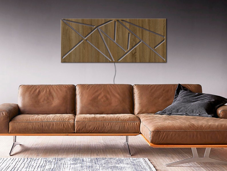 May include: A modern wooden wall sculpture with a geometric design. The sculpture is lit from behind, creating a warm glow. The sculpture is mounted on a gray wall above a brown leather sectional sofa.