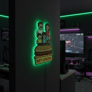 May include: A pixelated LED sign featuring the Bob's Burgers family atop a burger, with the text "BOB'S BURGERS" on a sign below. The sign has a green neon glow and is mounted on a dark wall.