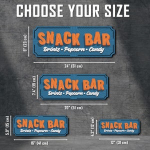 May include: Four different sizes of a blue and orange "Snack Bar" sign with the text "Drinks - Popcorn - Candy". The signs are shown against a gray textured background with measurements in inches and centimeters.