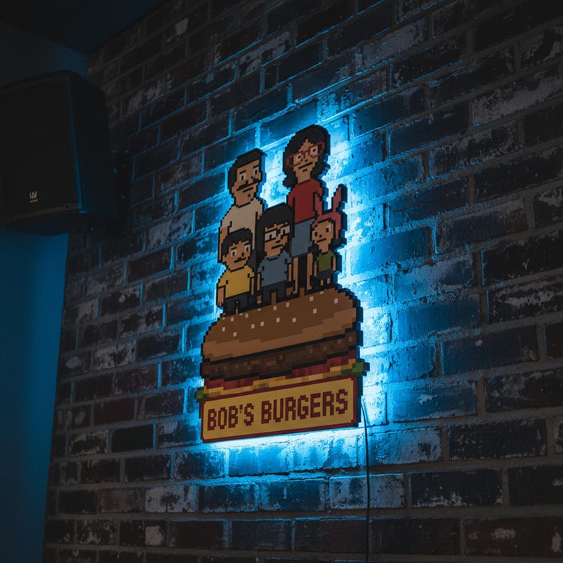 May include: Pixelated LED sign featuring the Bob's Burgers family atop a burger. The sign is mounted on a brick wall and illuminated with blue light. The sign reads "BOB'S BURGERS" in yellow letters.