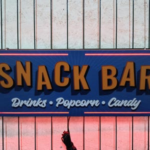 May include: A blue and orange sign with the text "SNACK BAR" in large letters and "Drinks - Popcorn - Candy" in smaller letters. The sign has a red neon outline.