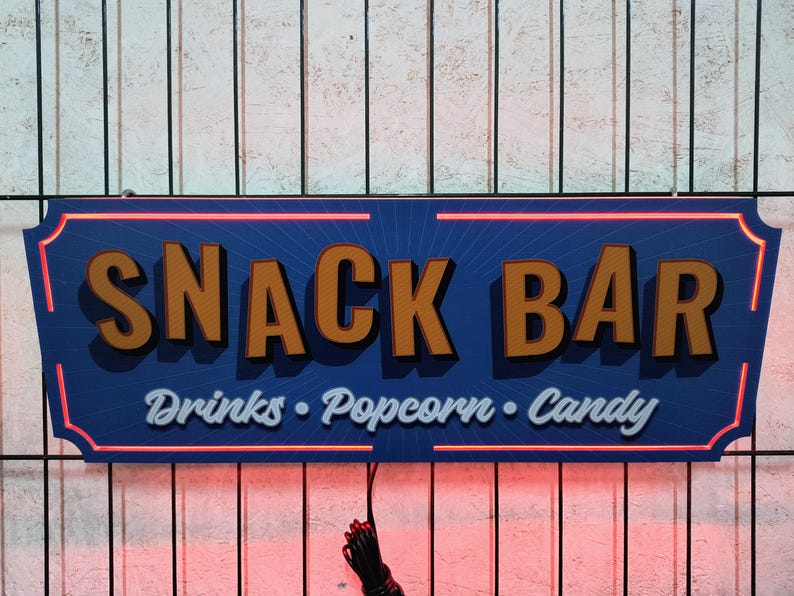 May include: A blue and orange sign with the text "SNACK BAR" in large letters and "Drinks - Popcorn - Candy" in smaller letters. The sign has a red neon outline.