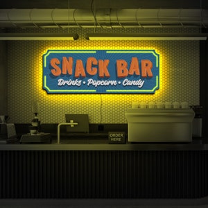 May include: A retro-style snack bar sign with yellow neon lights. The sign reads "Snack Bar" with the words "Drinks, Popcorn, Candy" underneath. The sign is mounted on a wall with a honeycomb pattern.