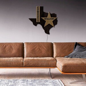 May include: A wooden Texas state outline with a gold star and the word "WILLIAM" inside the star. The word "COWBOYS" is written vertically on the left side of the outline. The outline is mounted on a wall above a brown leather sectional sofa.