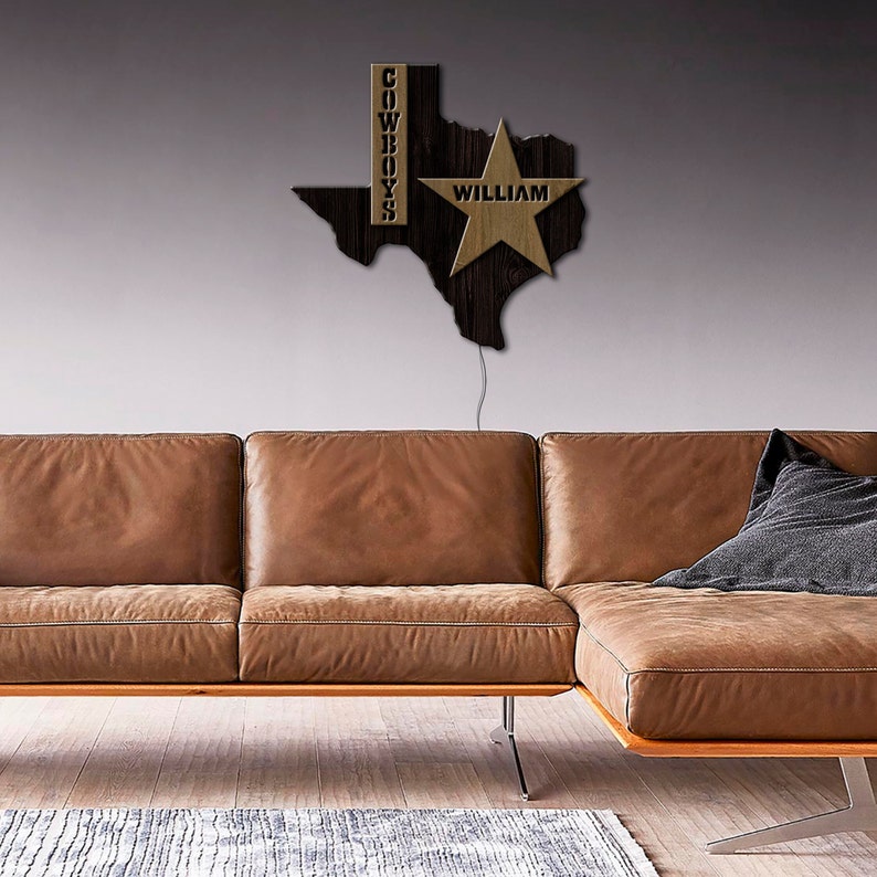 May include: A wooden Texas state outline with a gold star and the word "WILLIAM" inside the star. The word "COWBOYS" is written vertically on the left side of the outline. The outline is mounted on a wall above a brown leather sectional sofa.
