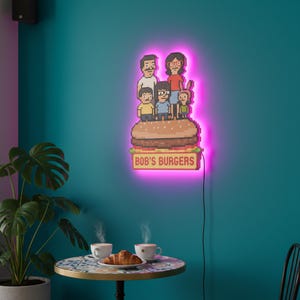 May include: A neon sign featuring the Bob's Burgers family atop a pixelated burger, with the text "BOB'S BURGERS" below. The sign has a bright pink glow and is mounted on a teal wall. A table with coffee and a croissant is in the foreground.