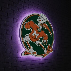 May include: A circular sign featuring the University of Miami Hurricanes mascot, a stylized pelican in an orange jersey with the letter "M". The sign is surrounded by a green and white border and is illuminated with a purple glow against a brick wall.