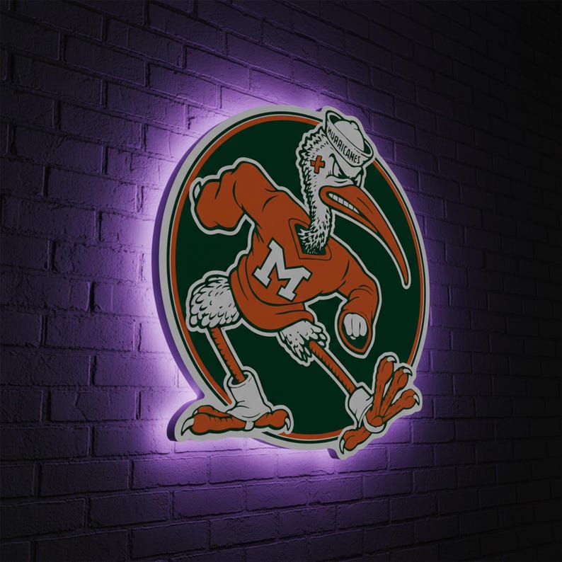 May include: A circular sign featuring the University of Miami Hurricanes mascot, a stylized pelican in an orange jersey with the letter "M". The sign is surrounded by a green and white border and is illuminated with a purple glow against a brick wall.