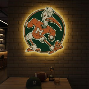 May include: A lighted sign featuring a cartoon pelican in an orange sweater with a white "M" on a green circle. The pelican wears a sailor hat. A football, trophy, hat, and glass of liquid sit on a wooden table in front of a brick wall.