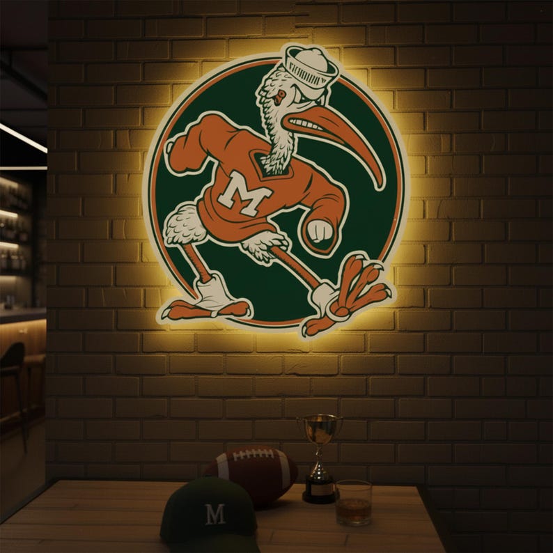 May include: A lighted sign featuring a cartoon pelican in an orange sweater with a white "M" on a green circle. The pelican wears a sailor hat. A football, trophy, hat, and glass of liquid sit on a wooden table in front of a brick wall.