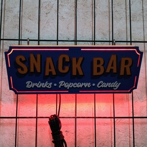 May include: A blue and orange sign with the text "Snack Bar" and the words "Drinks, Popcorn, Candy" written below. The sign is lit up with red lights.