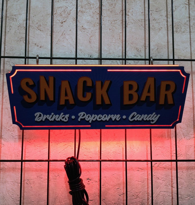 May include: A blue and orange sign with the text "Snack Bar" and the words "Drinks, Popcorn, Candy" written below. The sign is lit up with red lights.